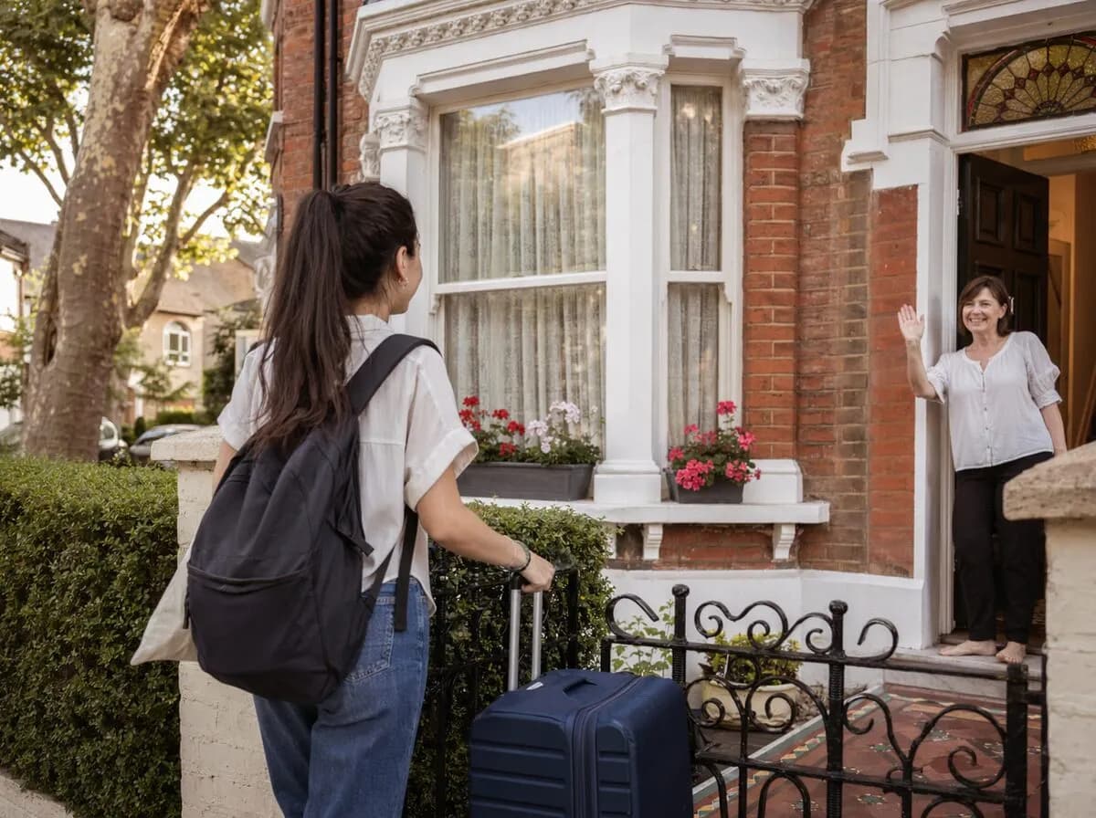 A guest arriving with a suitcase as their host waves from the doorway